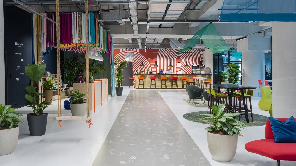 Blick durch den offenen Eingangsbereich auf das Café mit goldener Theke, Barhockern in Pink und Orange, Pflanzen und farbiger Deckendekoration.