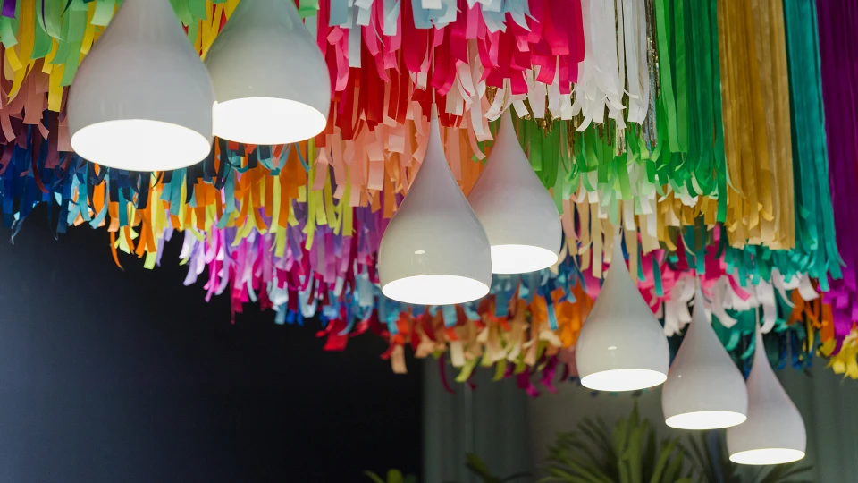 Weiße Pendelleuchten hängen unter einer farbenfrohen Deckeninstallation aus bunten Papierstreifen.
