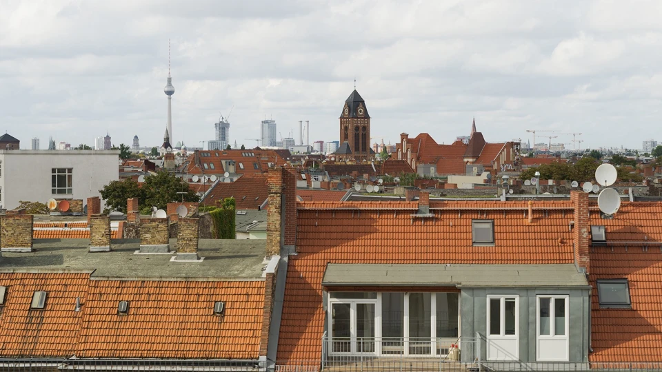 Stadtbild mit Dächern von Berlin, darunter rote Ziegeldächer und modernere Gebäude, im Hintergrund der Fernsehturm und eine Kirche.