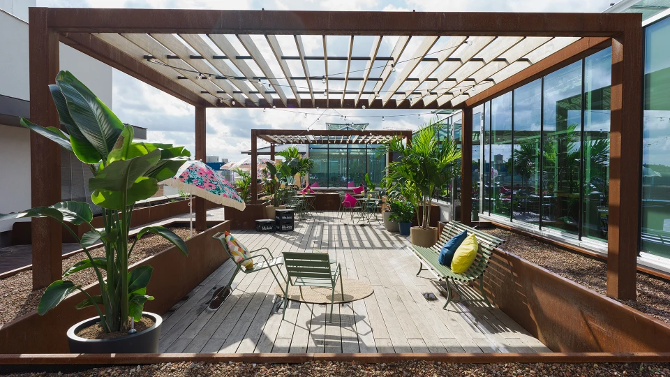 Modern gestaltete Dachterrasse mit Holzboden und -dach, bepflanzt mit großen Grünpflanzen und ausgestattet mit bunten Sitzmöbeln.