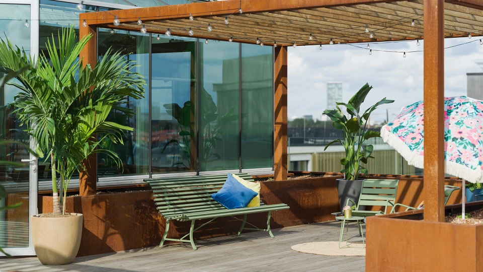 Gemütliche Terrasse eines modernen Gebäudes mit grünen Metallbänken, großen Topfpflanzen und einem bunten Sonnenschirm.