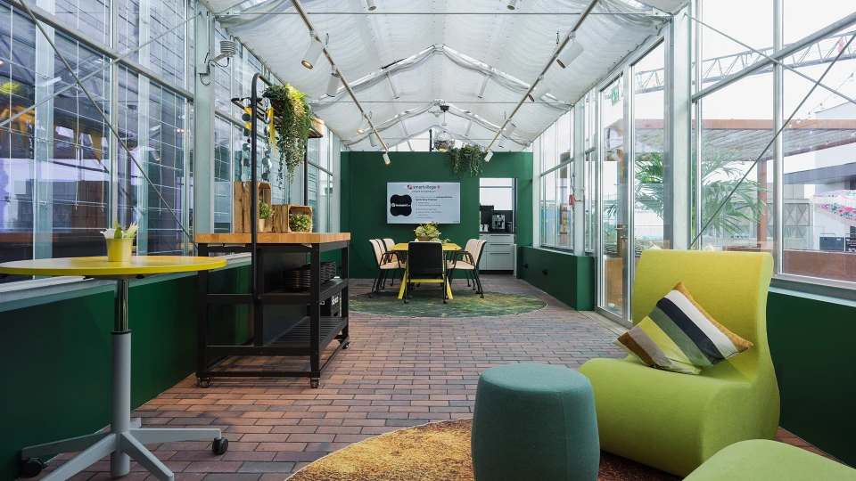 Moderner Bürobereich in einem gläsernen Gewächshaus mit grünen Wänden, Stühlen, Pflanzen und flexiblem Arbeitsbereich.