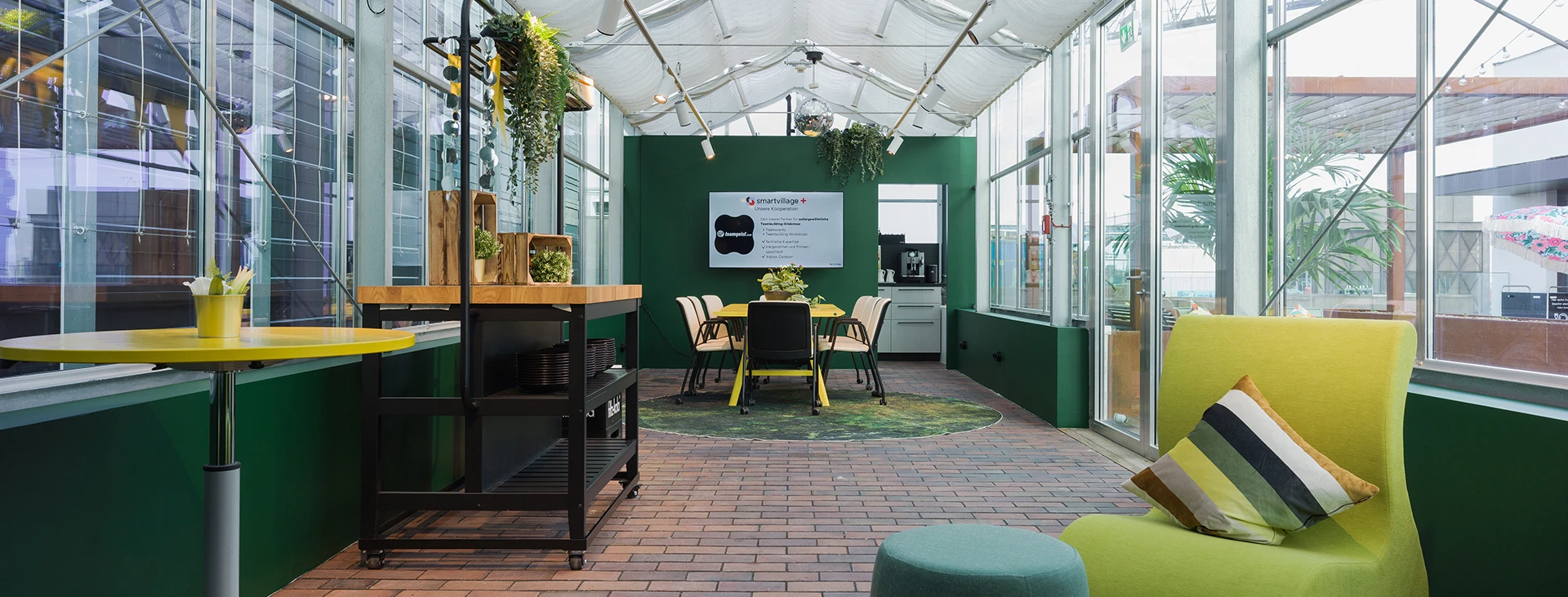 Moderner Bürobereich in einem gläsernen Gewächshaus mit grünen Wänden, Stühlen, Pflanzen und flexiblem Arbeitsbereich.
