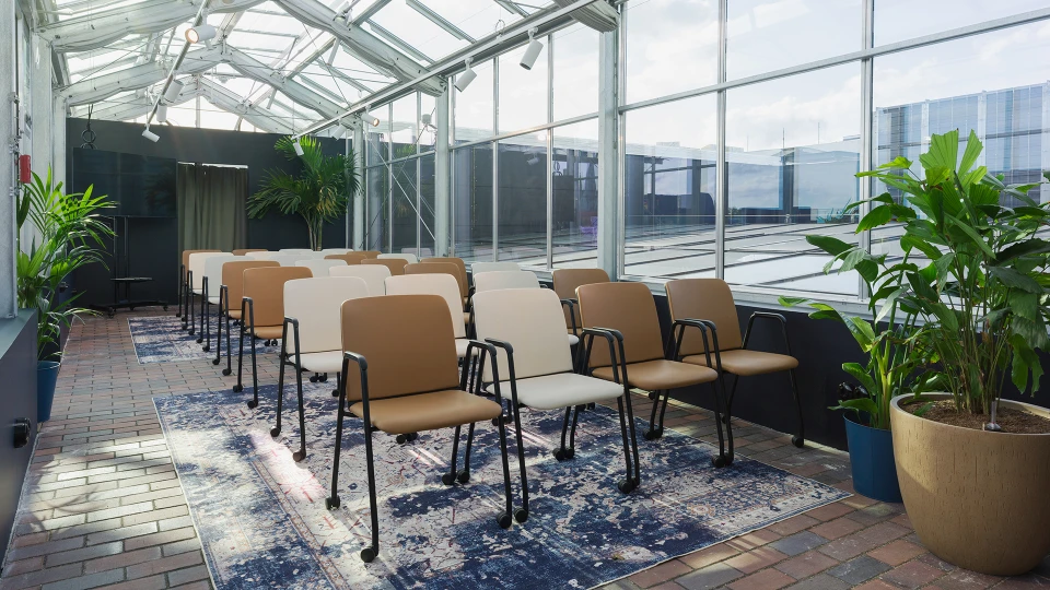 Ein heller, verglaster Raum mit einer Reihe beige Stühle auf einem blauen Teppich, umgeben von Pflanzen und Ziegelpflaster.