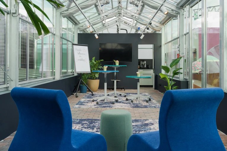 Modern eingerichtetes Büro in einem Glashaus mit zwei blauen Sesseln, einem höhenverstellbaren Schreibtisch, einem großen Bildschirm und einer Whiteboard-Tafel.