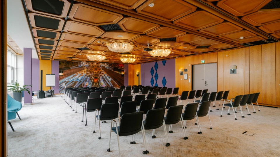 Eleganter Konferenzraum mit einer kunstvoll gestalteten Holzdecke, funkelnden Kronleuchtern und einer auffälligen Wandinstallation. Die Reihenbestuhlung mit schwarzen Stühlen auf Rollen bietet Platz für eine große Anzahl von Teilnehmern. An der Stirnseite des Raumes befindet sich eine kreative Wandgestaltung mit futuristischen Elementen, die eine inspirierende Atmosphäre schafft. Große Fenster auf der linken Seite sorgen für viel Tageslicht, während moderne Akzente in Lila und Blau dem Raum eine besondere Note verleihen