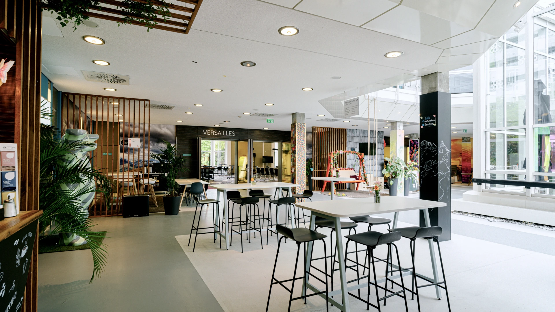 Modern gestalteter Lounge- und Empfangsbereich mit hohen Barhockern und Tischen in einem lichtdurchfluteten Raum. Im Hintergrund ist der Konferenzraum 'Versailles' mit geöffneter Glastür sichtbar, bereit für Veranstaltungen oder Meetings. Grüne Pflanzen und dekorative Holzelemente verleihen dem Ambiente eine natürliche und stilvolle Atmosphäre. Durch die großen Fenster gelangt viel Tageslicht in den offenen Bereich, der sich ideal für Networking und entspannte Gespräche eignet.
