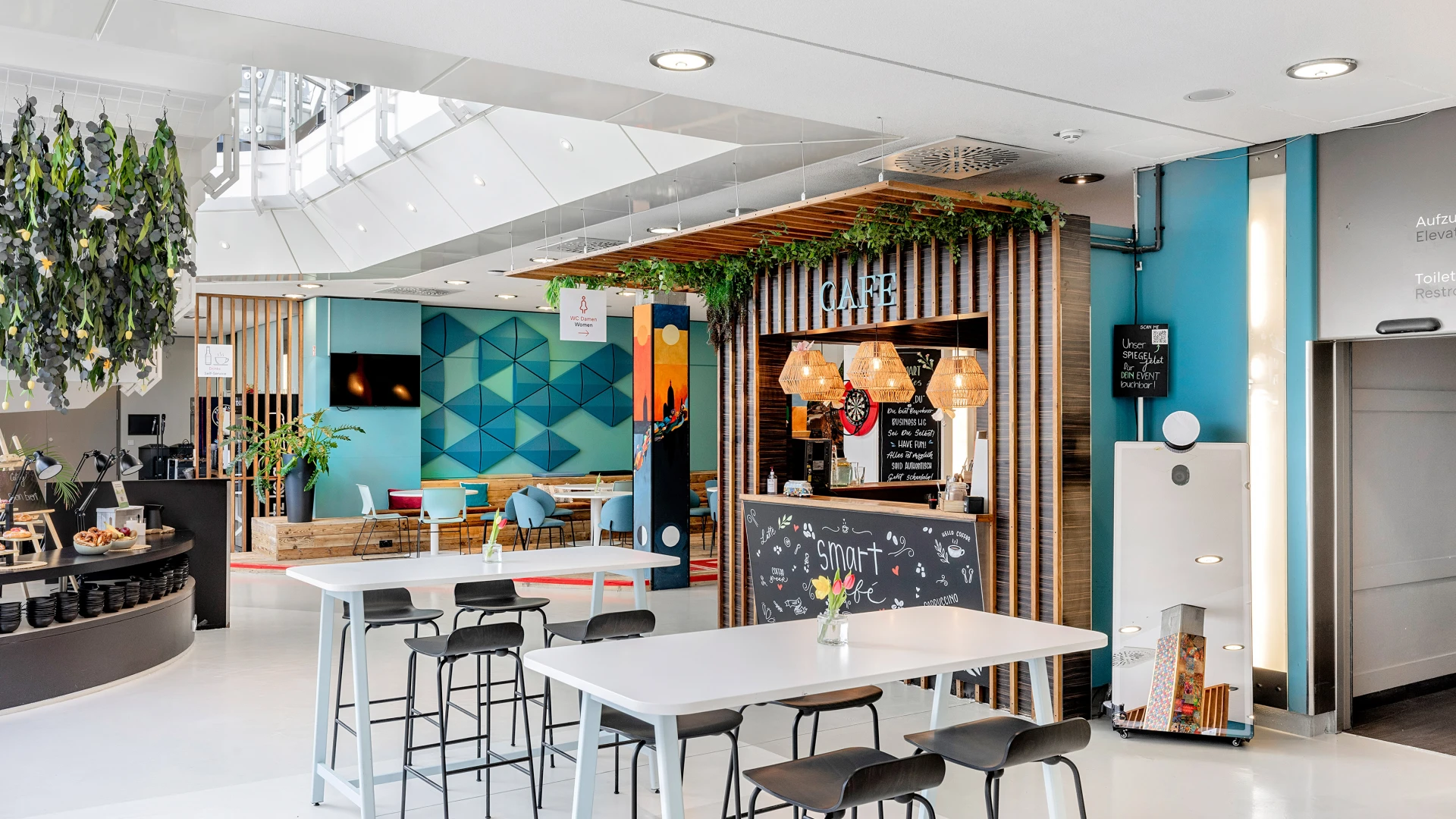 Helles, modernes Café mit weißen Tischen und schwarzen Barhockern, einer farbenfrohen Wandgestaltung und begrünten Holzelementen. Im Hintergrund befindet sich der Cafébereich mit Theke und Sitzplätzen.