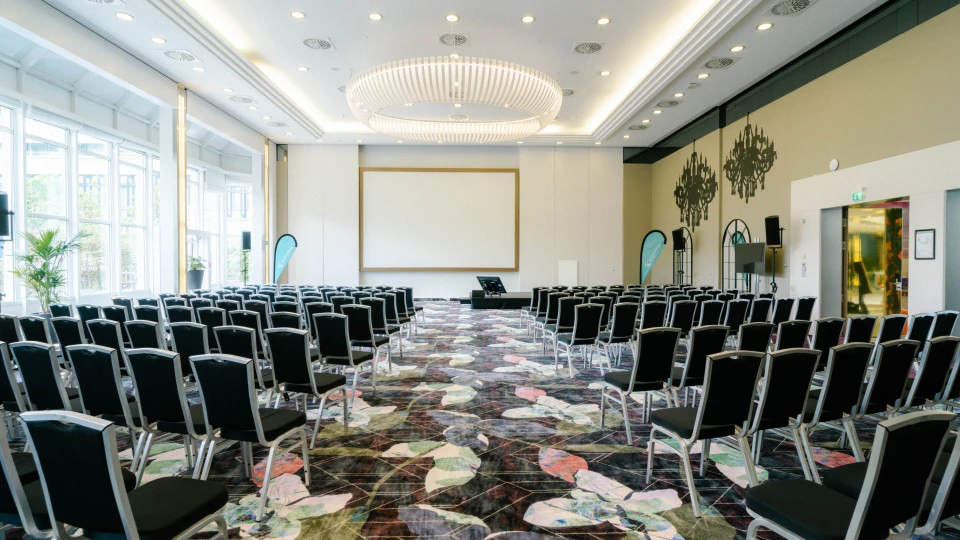 Symmetrischer Blick auf den Konferenzraum Wien mit zentraler Projektionsfläche, LED-Kronleuchter und großer Fensterfront.