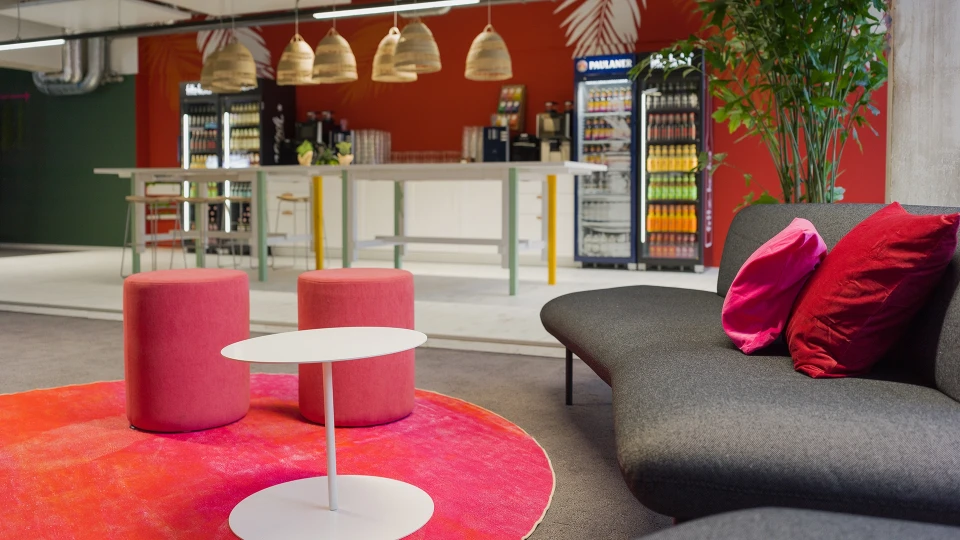 Sitzbereich mit dunklen Sofas, pinken Kissen und runden Hockern vor bodentiefen Fenstern im Smartvillage Köln-Mülheim.