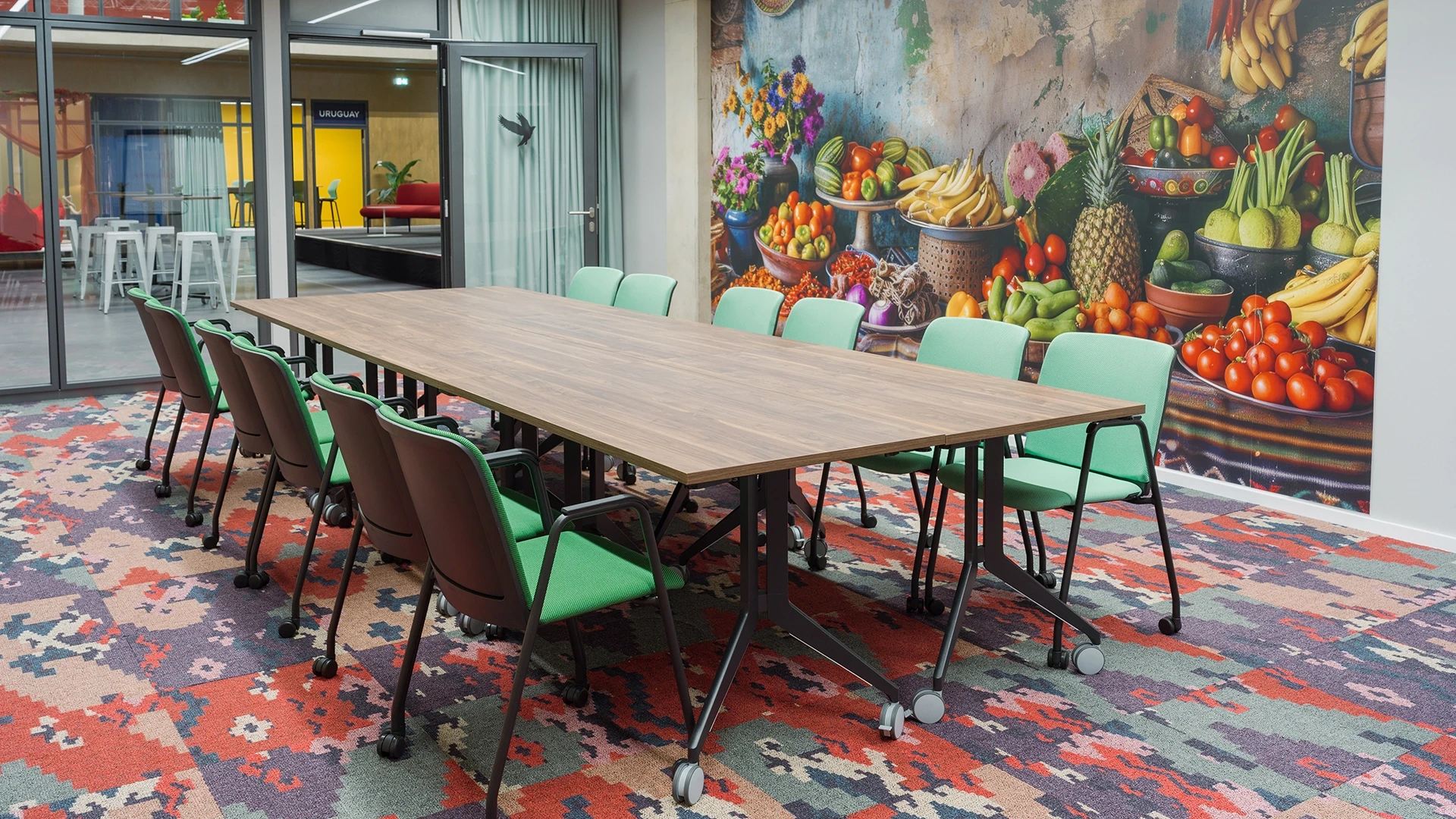 Konferenzraum Kolumbien Bogota mit großem Holztisch, grünen Stühlen und Wandbild eines Obst- und Gemüsemarkts, Blick auf Uruguay-Bereich im Smartvillage