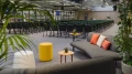Moderne Bürolounge mit gemütlichen Sofas und Kissen, einem gelben Hocker, kleinem Holztisch und vielen Grünpflanzen, dahinter ein großer Konferenzraum mit vielen Stühlen.