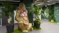Eine goldene Gorilla-Skulptur sitzt in einem Büroflur, geschmückt mit einer pinken Blumenkette, umgeben von mehreren großen Grünpflanzen.