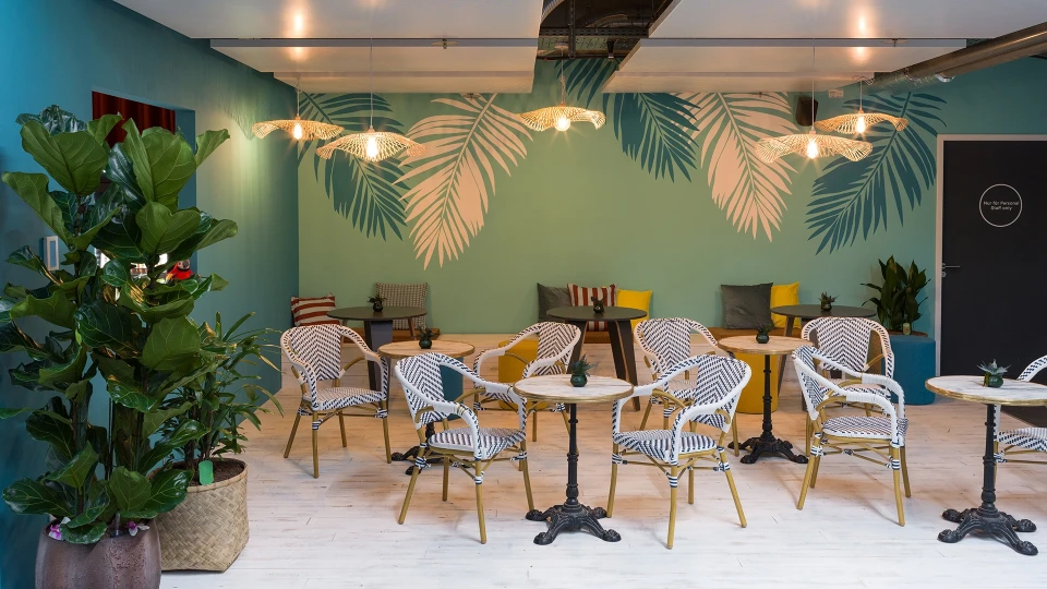 Stilvoller Innenraum eines Cafés mit tropischem Thema, hellen Wandfarben, gemusterten Stühlen und einer großen Pflanze links