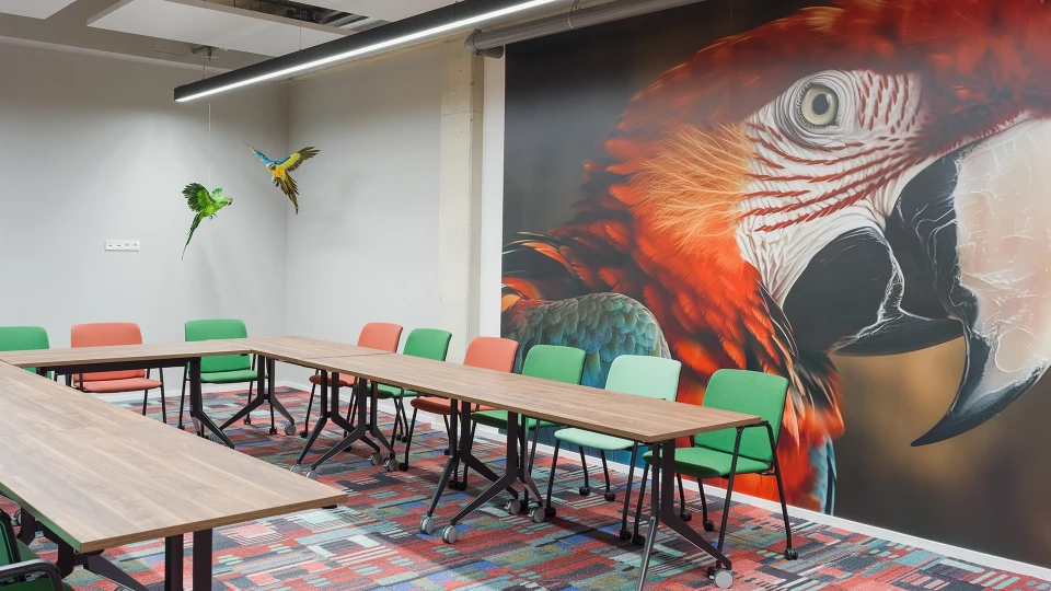 Ein moderner Besprechungsraum mit einem großen Tisch und mehreren Stühlen. An der Wand ist ein großes, farbenfrohes Wandgemälde eines Papageienkopfes.