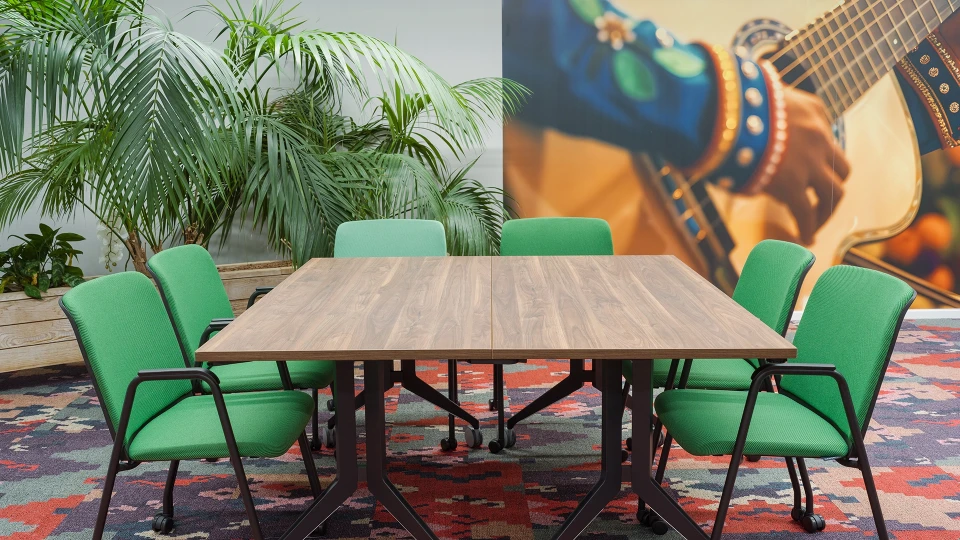 Ein modernes Büro mit einem großen Holztisch, umgeben von grünen Stühlen auf einem bunten Teppich, neben einer Wand mit Palmen und einem Poster von einer Gitarre.