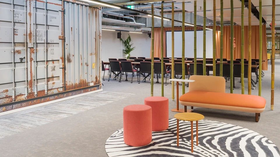 Modernes Büro mit einer Kombination aus rustikalen Containerwänden und stilvollen Möbeln, darunter ein orangefarbenes Sofa, runde rosa Hocker und ein schwarz-weißer Teppich.
