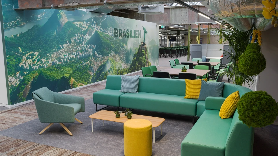Modernes Büro-Lounge mit grünen Sofas, Sesseln und gelben Akzenten vor einer großen Wandgrafik mit der Aufschrift 'Lebe Brasilien'