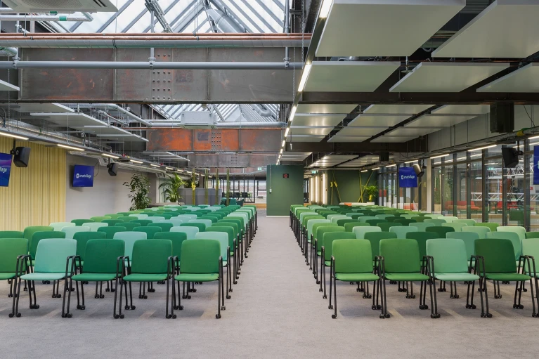 Leerer Konferenzraum mit Reihen von grünen Stühlen, beleuchtet durch Oberlichter, umgeben von industriellen Metallstrukturen und Glaswänden. An den Wänden hängen Monitore mit Logos.