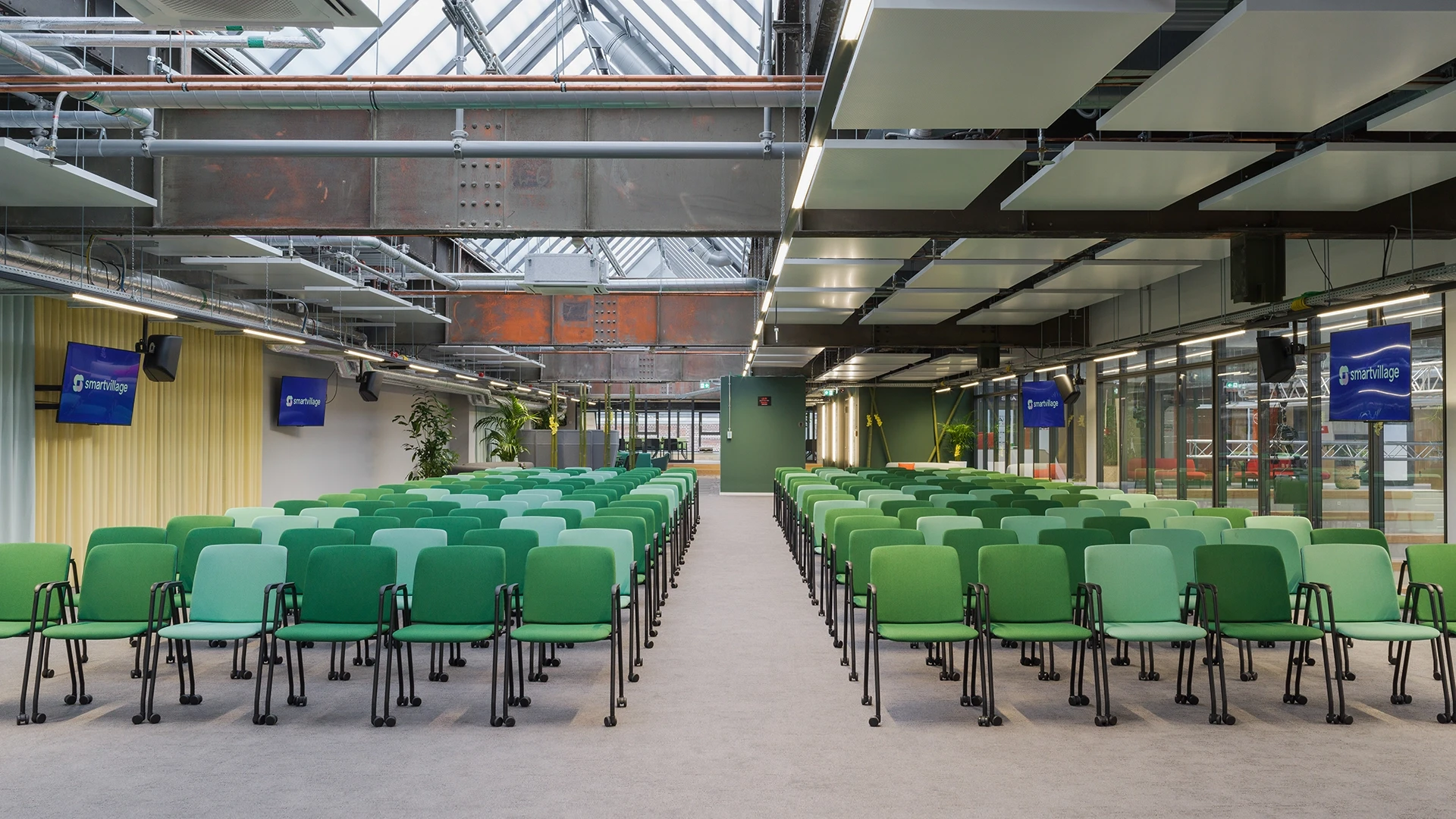 Leerer Konferenzraum mit Reihen von grünen Stühlen, beleuchtet durch Oberlichter, umgeben von industriellen Metallstrukturen und Glaswänden. An den Wänden hängen Monitore mit Logos.