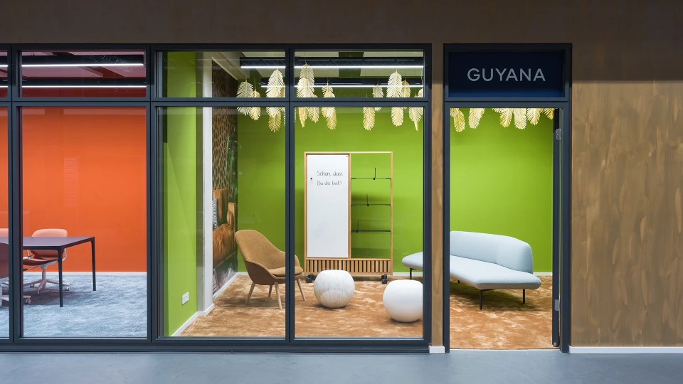 Innenaufnahme des Raumes "Guyana" mit beigem Teppichboden, hellem Sofa, braunem Loungesessel und weißem Kugelhocker vor grünem Hintergrund.