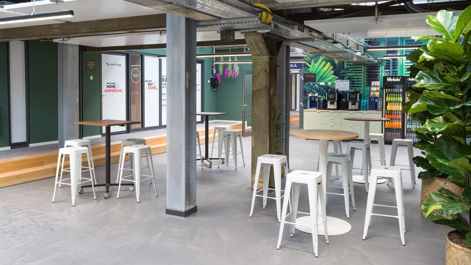 Raum mit Stehtischen und weißen Barhockern im Smartvillage Köln Mülheim Jungle Lodge Bereich.