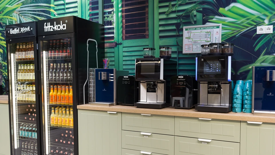 Sitzbereich mit Hochtisch und Barhockern vor orangenen Vorhängen und großen Pflanzen.