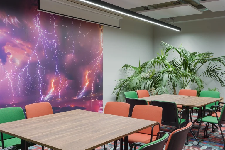 Besprechungsraum mit mehreren Tischen und Stühlen in Grün und Orange. Eine Wand ist mit einem großen Wandbild eines dramatischen lila Himmels mit Blitzen verziert.