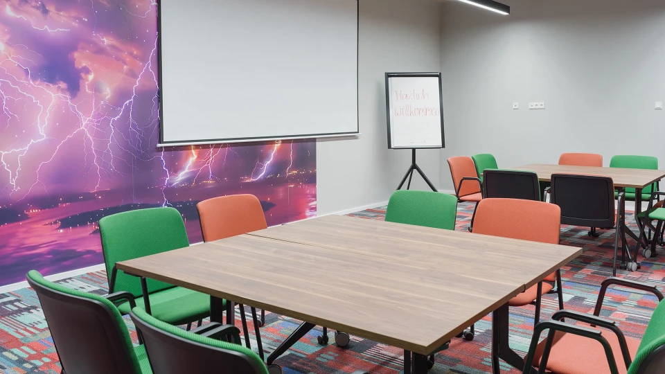 Modernes Schulungsraum-Interieur mit bunten Stühlen um Holztische, eine weiße Tafel und eine Wand mit einem leuchtenden Weltraumbild.
