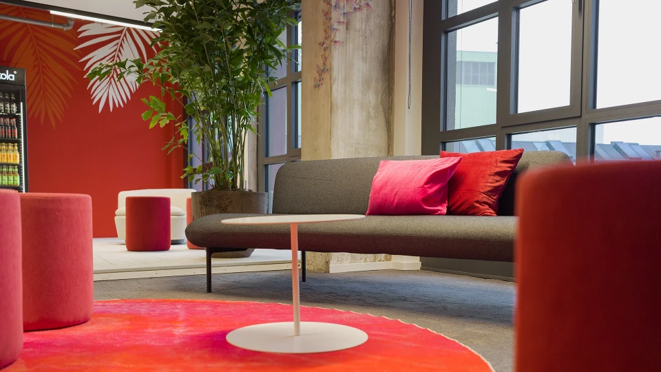 Sicht auf kleine Büro-Kabine mit Glastür vor dunkelgrüner Wand und floralem Dekor, im Hintergrund Lounge mit tropischem Strandmotiv.