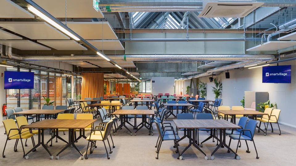 Weitwinkelaufnahme des Veranstaltungsbereichs mit vielen bunt gemischten Stühlen, Tischen und großem "smartvillage"-Monitor.