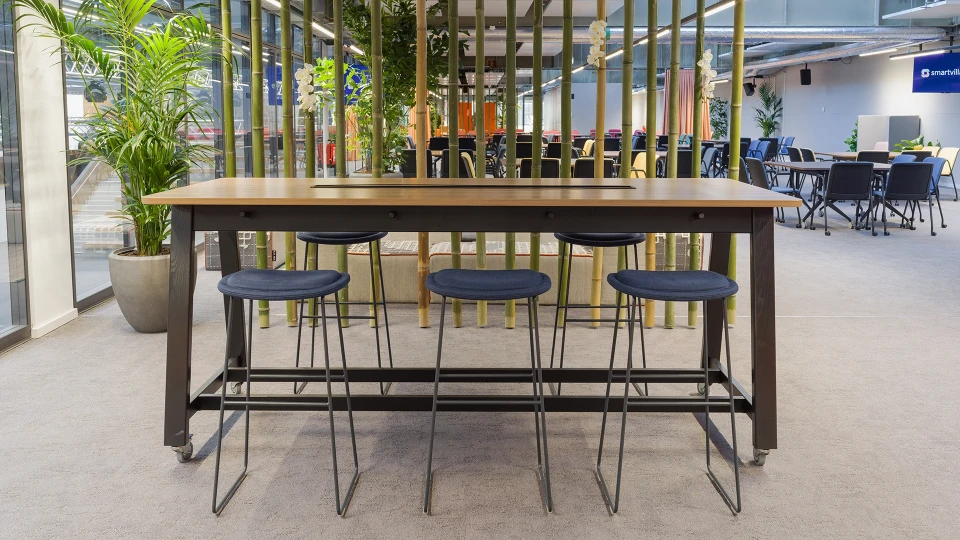 Ansicht eines großen Bartisches mit blauen Hockern vor Bambusdeko und Blick auf die offene Eventfläche im Hintergrund.