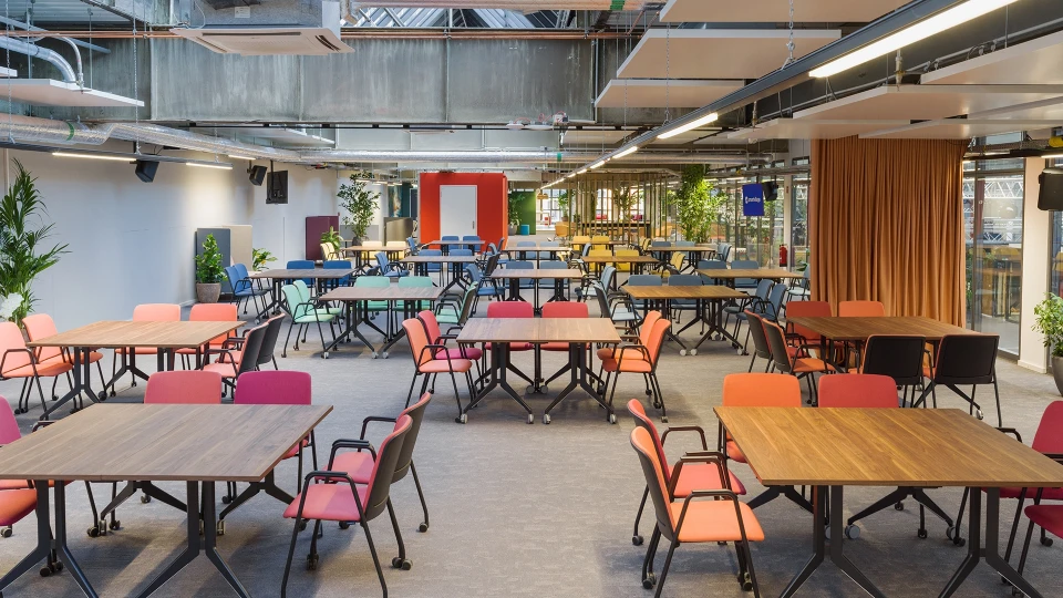 Große offene Fläche mit bunten Stühlen, Konferenztischen und urban-industriellem Deckenlook.
