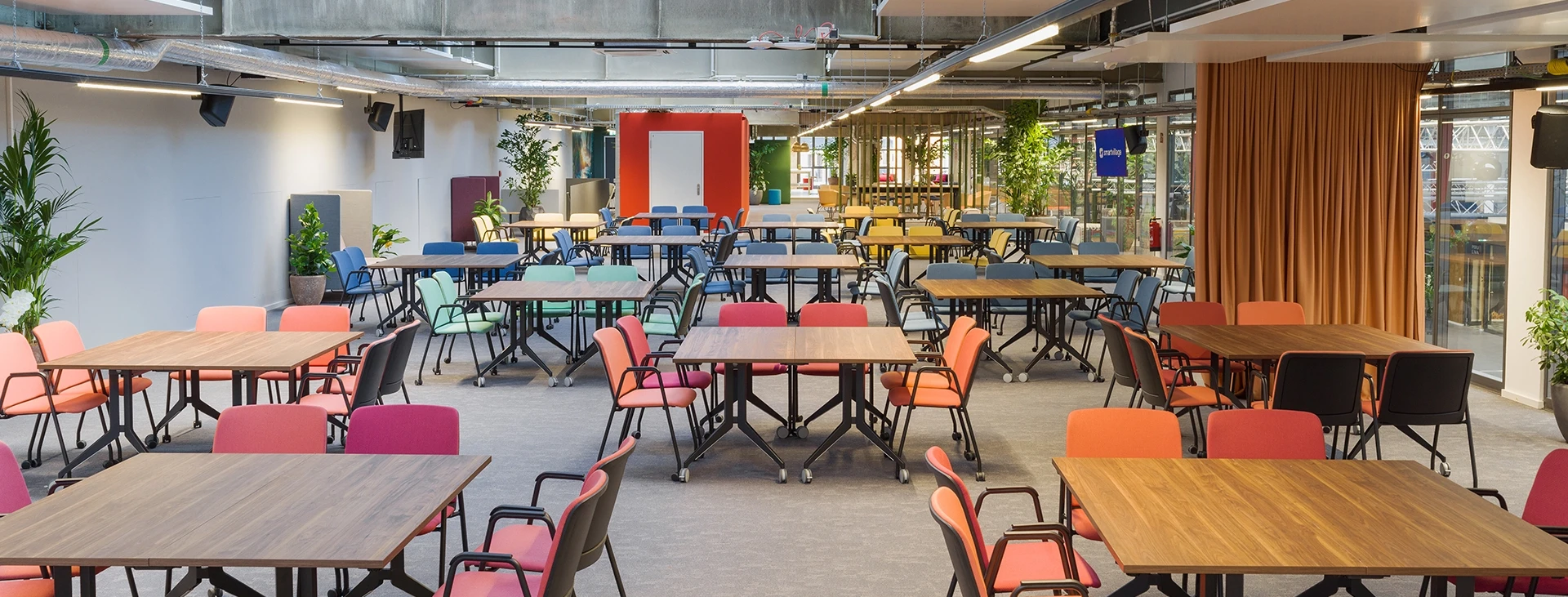 Große offene Fläche mit bunten Stühlen, Konferenztischen und urban-industriellem Deckenlook.