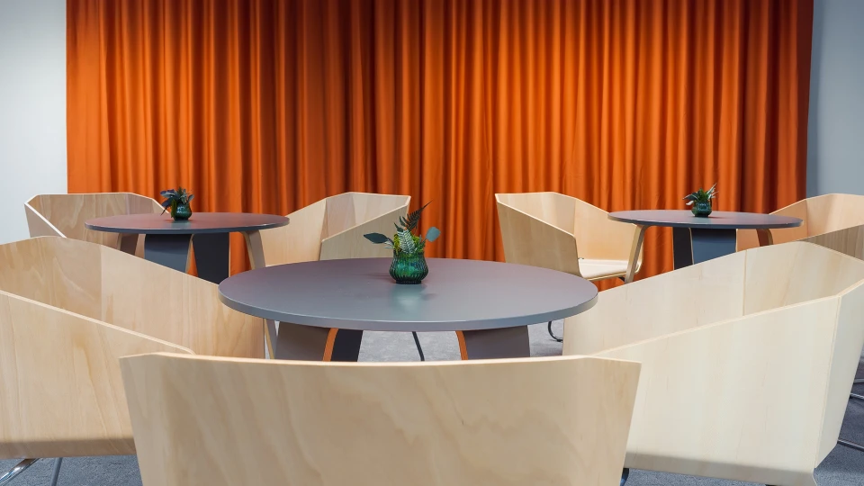 Kleine Bistro-Ecke mit runden Tischen und modernen Holzschalenstühlen vor einem orangefarbenen Vorhang.