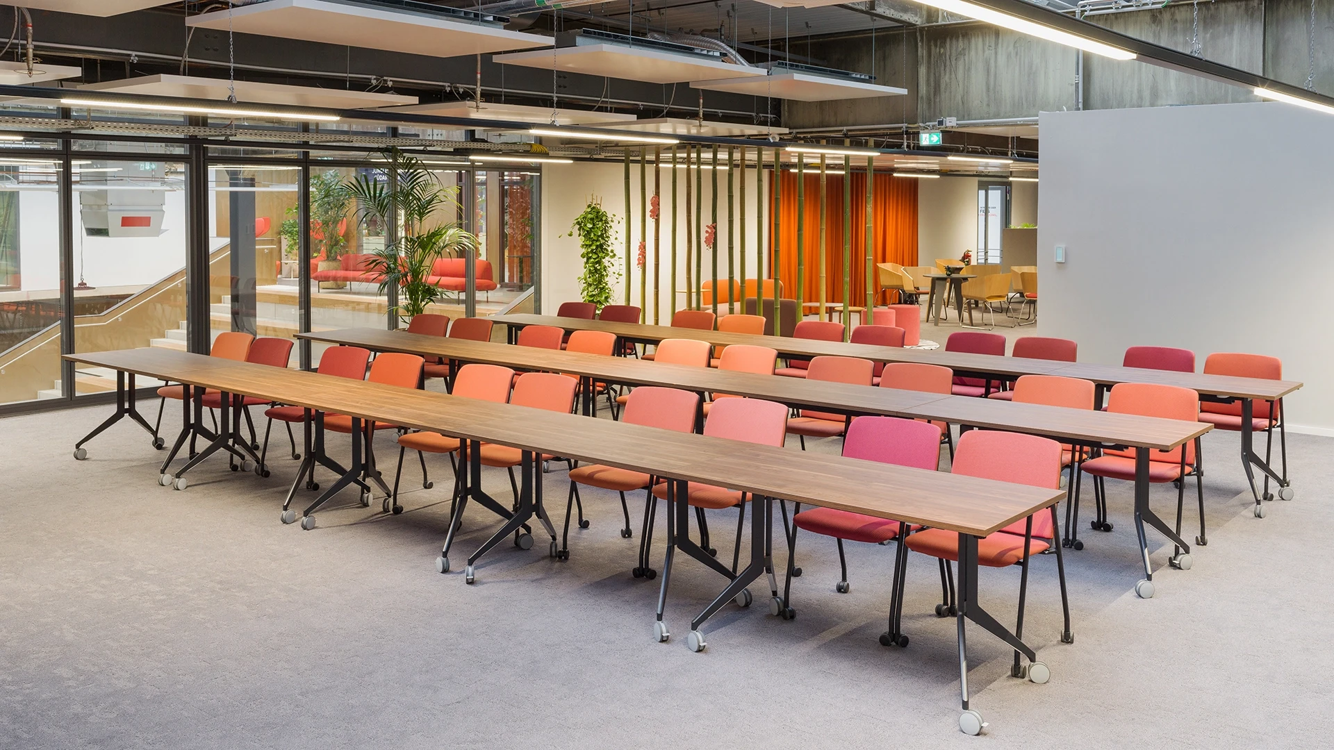 Seminarraum mit mehreren Reihen rollbarer Holztische und farblich gemischter Bestuhlung, im Hintergrund ein heller Loungebereich mit Bambus-Trennung.