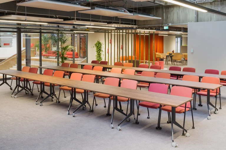 Seminarraum mit mehreren Reihen rollbarer Holztische und farblich gemischter Bestuhlung, im Hintergrund ein heller Loungebereich mit Bambus-Trennung.