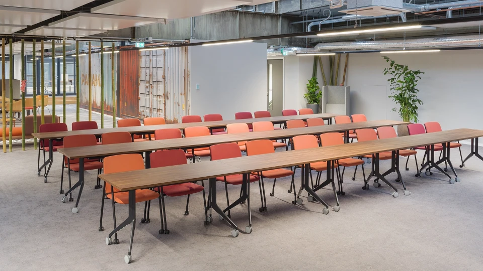 Seminarbereich mit drei Reihen rollbarer Tische und orangerot gemischter Bestuhlung vor weißer Wand und Industrie-Decke, im Hintergrund ein Container mit "PANAMA"-Schriftzug.