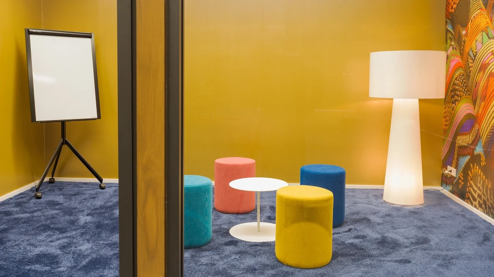 Nahaufnahme von fünf farbigen Poufs (gelb, türkis, blau, orange, pink) um einen runden weißen Beistelltisch auf dunklem Teppichboden.