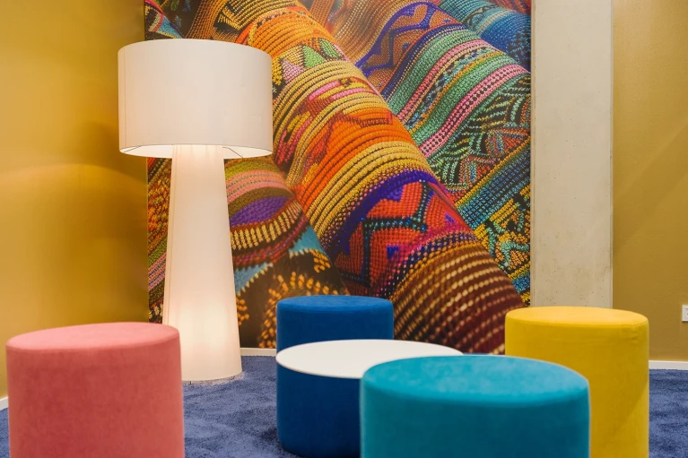 Innenansicht des Paraguay-Raums mit großer Stehlampe, farbigen Sitzhockern und einer detailreichen Wandgrafik mit buntem Textilgewebe.