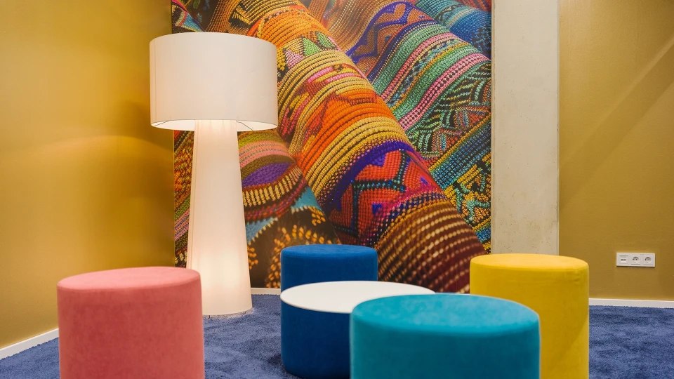 Innenansicht des Paraguay-Raums mit großer Stehlampe, farbigen Sitzhockern und einer detailreichen Wandgrafik mit buntem Textilgewebe.