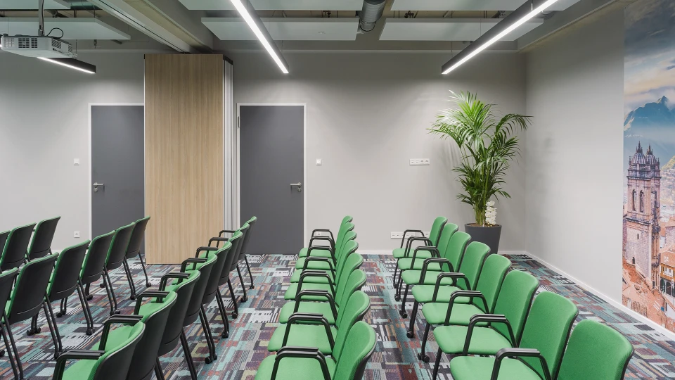 Seminarraum mit Leinwand und Reihen aus grünen Stühlen, geöffnet zur Caféecke.