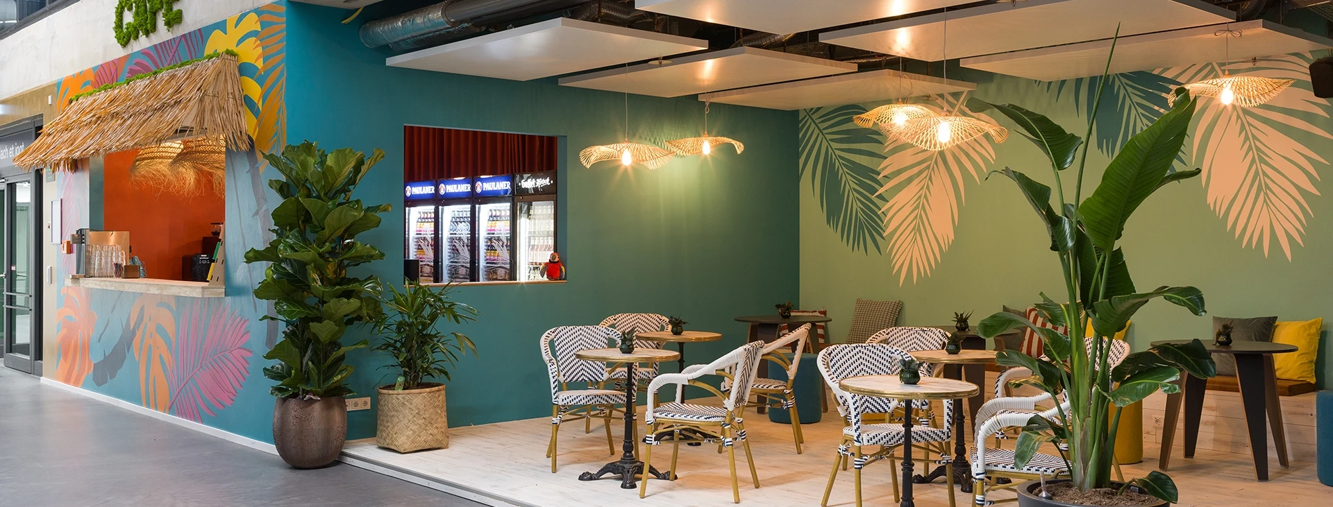 Frontalansicht des Smartcafés mit runder Café-Bestuhlung vor einer Wand mit großen Palmenmotiven.