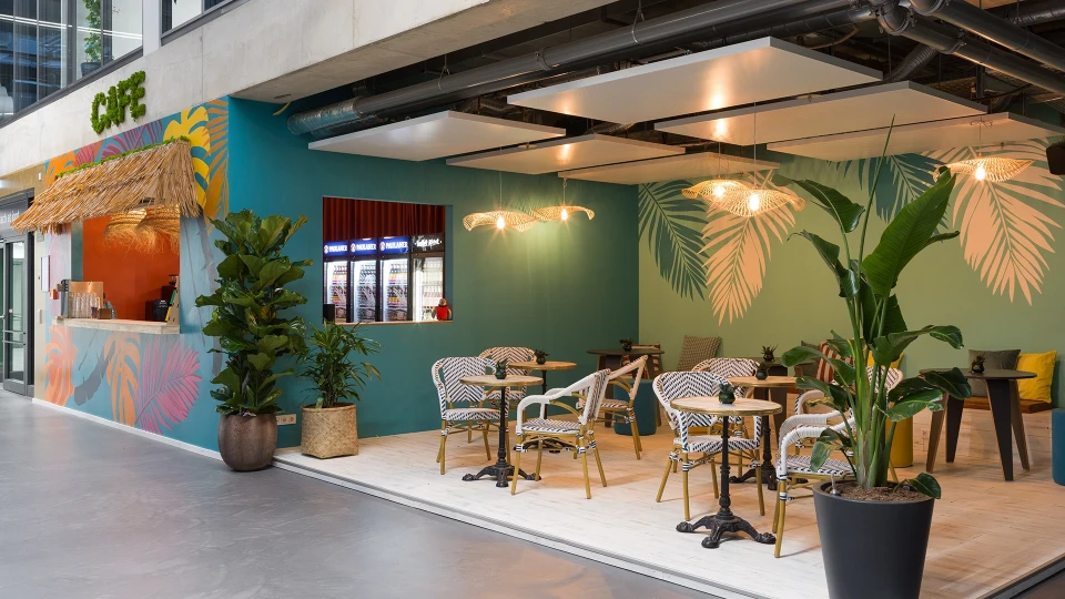 Frontalansicht des Smartcafés mit runder Café-Bestuhlung vor einer Wand mit großen Palmenmotiven.