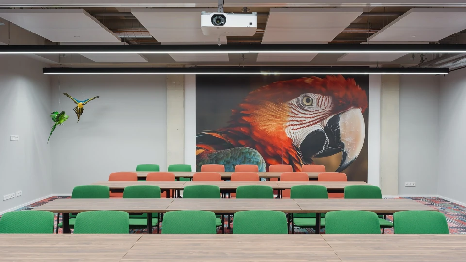 Besprechungsraum mit einem großen Tisch und grünen Stühlen, an der Wand ein großes Bild eines Papageienkopfes und eine kleine Skulptur eines fliegenden Vogels.
