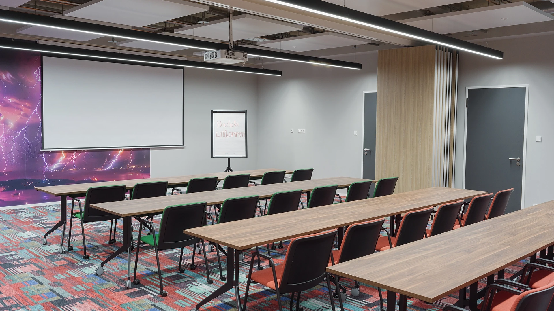 Modern conference room with long tables and black chairs, a white screen at the front, and a colorful space-themed wall