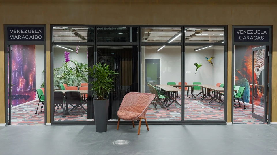 Moderne Bürolobby mit Glastüren, beschriftet mit 'Venezuela Maracaibo' und 'Venezuela Caracas', einladendem Innenraum mit bunten Stühlen und Pflanzen
