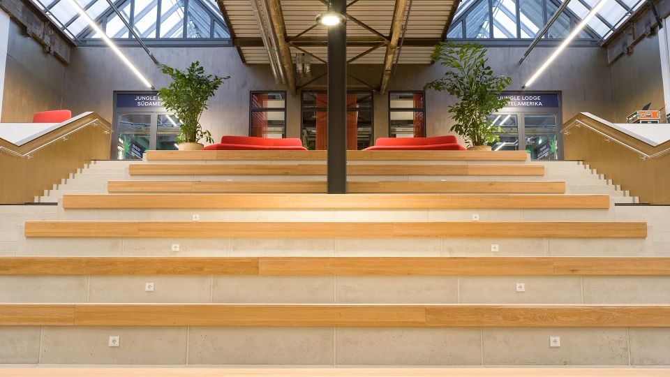 Moderne Innentreppe aus hellem Holz mit Stufen, die als Sitzgelegenheiten mit roten Kissen dienen, in einer offenen Halle mit Industriedesign.