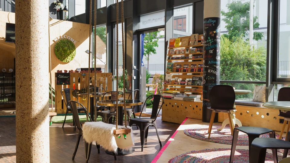 Innenansicht eines gemütlichen Cafés mit vielfältigen Sitzmöglichkeiten, darunter Stühle mit Fellbezug, und einem Regal voller Produkte zur Selbstbedienung am Tageslicht.