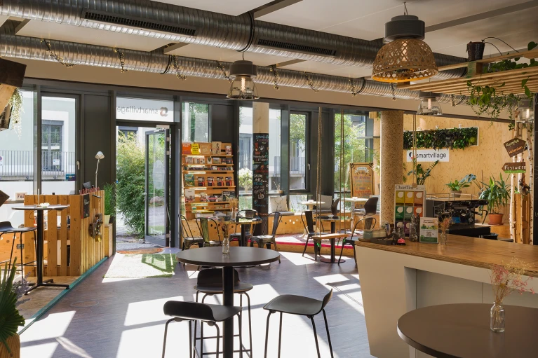 Heller, modern eingerichteter Café-Innenraum mit verschiedenen Sitzbereichen, Bartheke und Pflanzen. Blick durch große Fenster auf eine Terrasse.